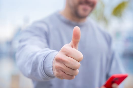 Unrecognizable portrait individuality Caucasian man outdoors near the water smiles and gives a thumbs up while holding a smartphone.His casual look and cheerful gesture express positivity and approval