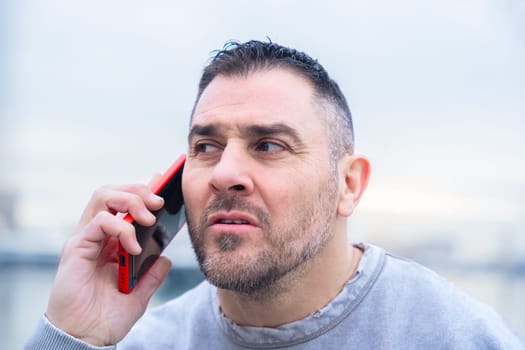 Caucasian man stands outdoors near water, talking on a smartphone with a serious look. His casual outfit and intense expression suggest concentration during an important phone conversation.