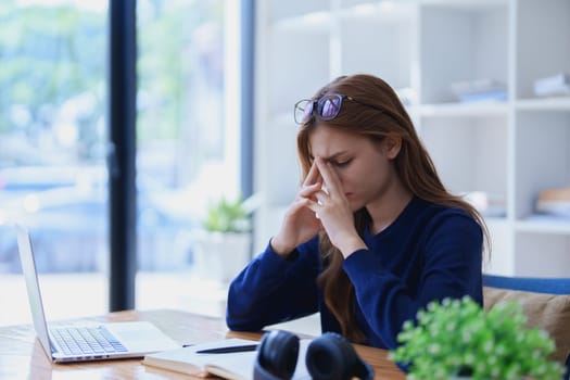 Stressed young woman overwhelmed by e-learning studies at desk with laptop, notebook, headphones, hands clasped to her face in frustration.