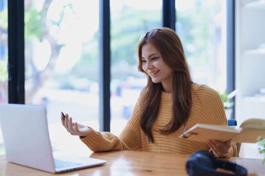 Smiling young woman engaged in a joyful online e-learning video call, gesturing while holding a book with headphones on a wooden table nearby.