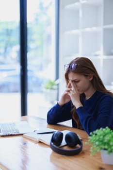 Stressed young woman overwhelmed by e-learning studies at desk with laptop, notebook, headphones, hands clasped to her face in frustration.