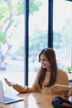 Smiling young woman engaged in a joyful online e-learning video call, gesturing while holding a book with headphones on a wooden table nearby.
