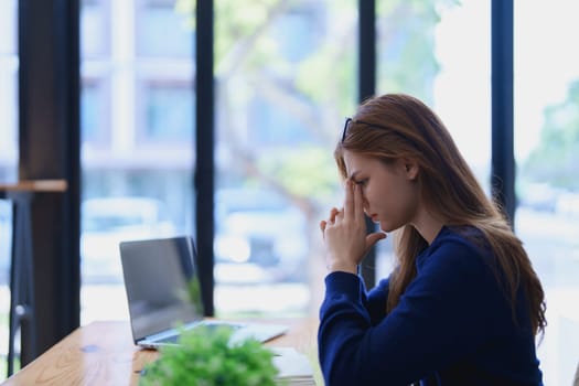Stressed young woman overwhelmed by e-learning studies at desk with laptop, notebook, headphones, hands clasped to her face in frustration.