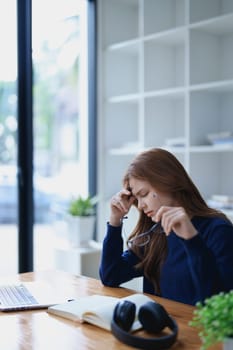 Stressed young woman overwhelmed by e-learning studies at desk with laptop, notebook, headphones, hands clasped to her face in frustration.