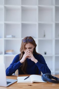 Stressed young woman overwhelmed by e-learning studies at desk with laptop, notebook, headphones, hands clasped to her face in frustration.