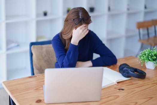 Stressed young woman overwhelmed by e-learning studies at desk with laptop, notebook, headphones, hands clasped to her face in frustration.