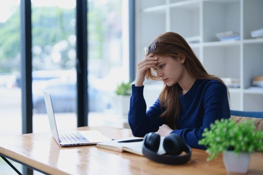 Stressed young woman overwhelmed by e-learning studies at desk with laptop, notebook, headphones, hands clasped to her face in frustration.