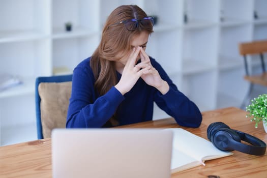 Stressed young woman overwhelmed by e-learning studies at desk with laptop, notebook, headphones, hands clasped to her face in frustration.