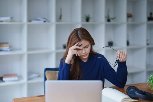 Stressed young woman overwhelmed by e-learning studies at desk with laptop, notebook, headphones, hands clasped to her face in frustration.