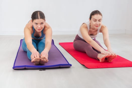 Caucasian women doing forward bend stretches on yoga mats in a bright indoor space. Ideal for fitness, wellness, flexibility, and healthy lifestyle concepts focused on movement and balance.
