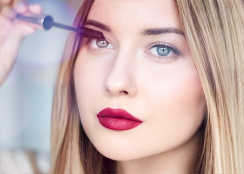 Woman applying mascara with confidence, defined brows and red lipstick. Blonde beauty focused on her eye makeup, cosmetics look
