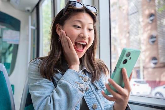Chinese woman reacts with joyful surprise while looking at smartphone inside a tram. Wearing denim jacket and sunglasses, she enjoys a fun and unexpected moment during her urban commute