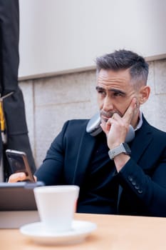 Caucasian mature man with headphones around neck looks thoughtful while checking smartphone. Seated at outdoor cafe with tablet and coffee, reflecting a modern and focused work lifestyle .Vertical