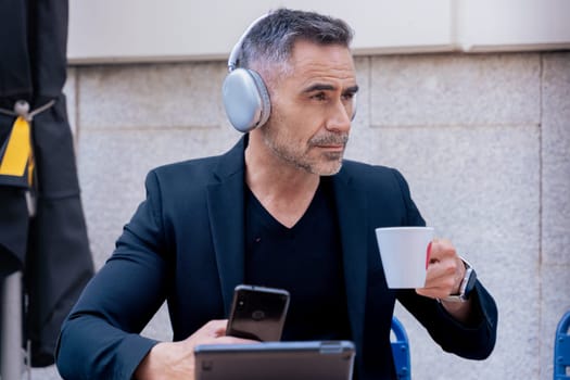 Caucasian mature man wearing headphones, holding smartphone and coffee while using tablet at an outdoor cafe. Multitasking with style, enjoying modern technology and a peaceful break