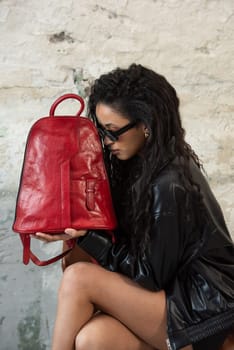 Curly girl wearing black leather jacket, sunglasses, short skirt posing with a red backpack.