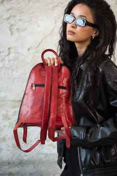 Curly girl wearing black leather jacket, sunglasses, shoes and short skirt posing with a red backpack.