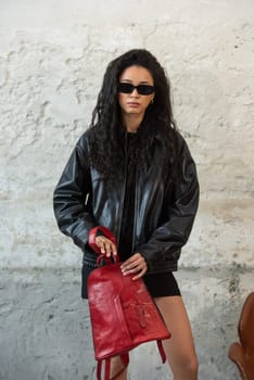 Curly girl wearing black leather jacket, sunglasses, shoes and short skirt posing with a red backpack.