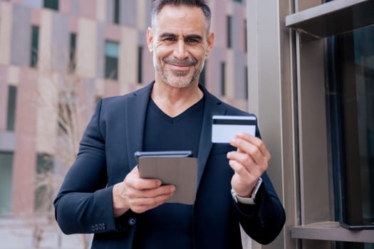 Caucasian mature man smiling at camera while holding a credit card and tablet. Standing outdoors in urban setting, representing modern technology, online shopping, and financial confidence
