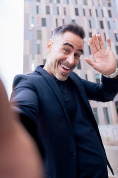 Caucasian mature man taking a cheerful selfie while waving at the camera. Dressed in black, smiling widely in an urban background, capturing a fun and spontaneous moment outdoors .Vertical