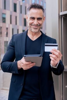 Caucasian mature man smiling at camera while holding a credit card and tablet. Standing outdoors in urban setting, representing modern technology, online shopping, and financial confidence .Vertical