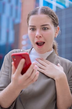 Caucasian woman looking at her smartphone with a shocked and surprised expression. Unexpected news, social media reaction, and technology concept in an urban environment with modern buildings.Vertical