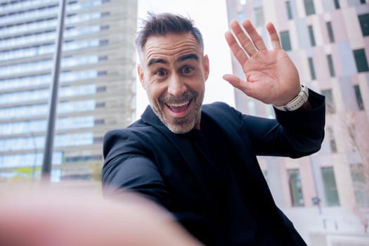 Caucasian mature man taking a cheerful selfie while waving at the camera. Dressed in black, smiling widely in an urban background, capturing a fun and spontaneous moment outdoors