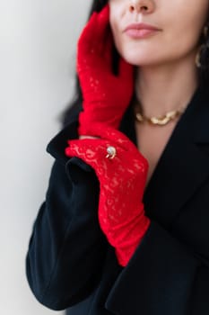 Elegant woman in red lace gloves and black blazer poses, touching face. Close-up highlights gold diamond ring over glove, gold necklace. Fashion, style, luxury accessories.