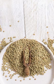Plate full of dried raw green lentils with a wooden scoop on white table top view, copy space. Healthy raw vegetarian food, legumes plant-based protein source. Traditional mediterranean ingredient