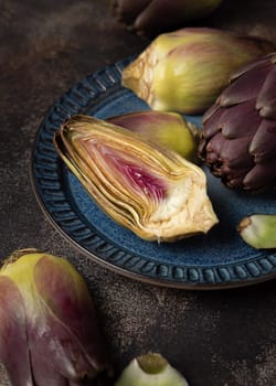 Blue plate full of purple raw romanesco artichokes on dark rustic table closeup. Fresh vegetables bought on farmers market ready to cook. Italian cousine ingredient