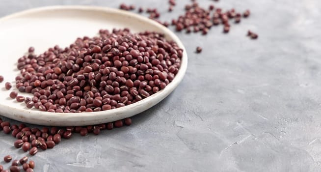 Ceramic plate full of dried red azuki beans on gray table closeup, copy space. Healthy vegetarian food, legumes protein source. Traditional East Asiancousin ingredient