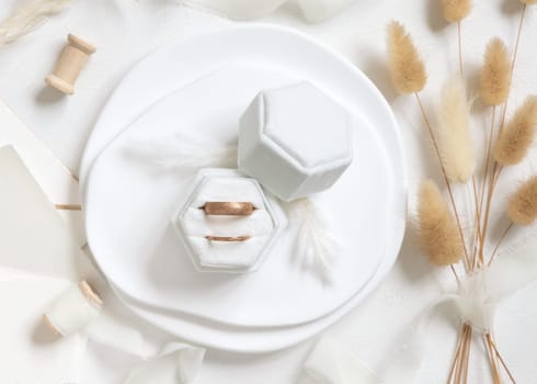 Golden wedding rings in a velvet jewelry box near silk ribbons and dried hare's tail grass top view on white. Boho romantic flatlay. Bohemian engagement proposal or Valentines day gift
