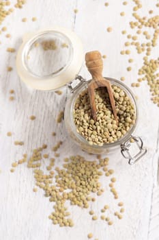 Opened glass jar full of dried green lentils with a wooden scoop on white table top view. Healthy vegetarian food, legumes plant-based protein source. Traditional mediterranean raw uncooked ingredient