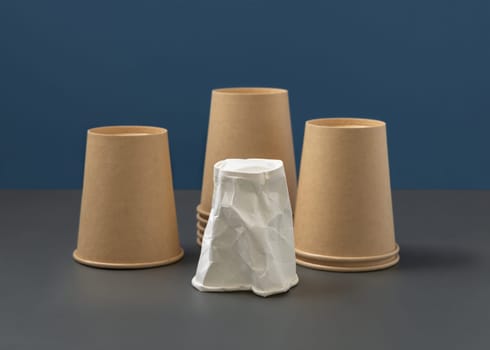 Disposable beige kraft paper cups and crumpled used white one on gray against blue closeup, mockup, copy space. Empty cardboard coffee cups. Consumerism concept