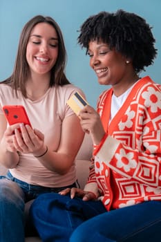 Cheerful caucasian and african american women sitting on couch enjoying a moment together while looking at a red smartphone, sharing laughter and spending quality time indoors .Vertical