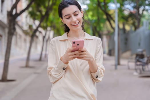 Caucasian woman smiling while using her smartphone on a quiet urban street. Casual summer outfit and relaxed atmosphere, surrounded by trees and buildings during a peaceful walk