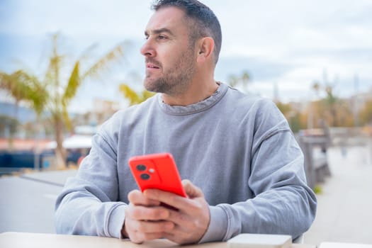 caucasian Man typing on a red smartphone, sitting outdoors by the port with palm trees, wearing a grey sweater, focused on the screen in a relaxed and casual atmosphere.