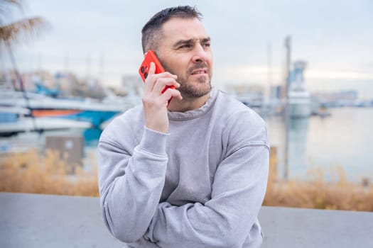 Caucasian man stands outdoors near water, talking on a smartphone with a serious look. His casual outfit and intense expression suggest concentration during an important phone conversation.