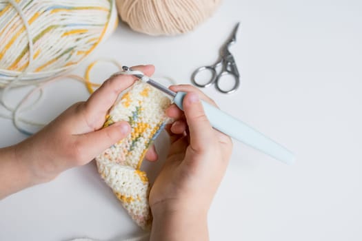 Child crocheting colorful yarn with hook and scissors on table.