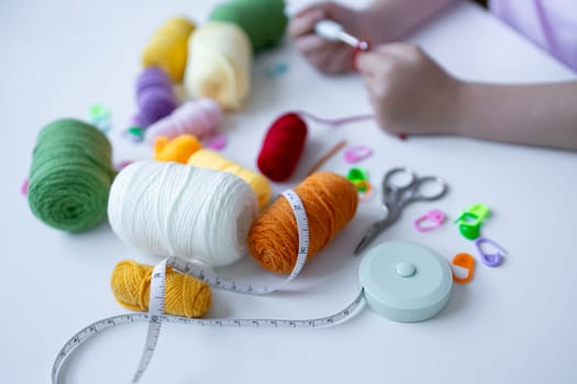 Young hands knitting with colorful yarn and measuring tape on white surface.