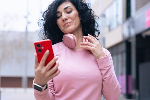 Caucasian mid adult woman with curly black hair standing in an urban environment. She wears a pink sweatshirt, pink headphones, and holds a smartphone, fitness, technology concept.