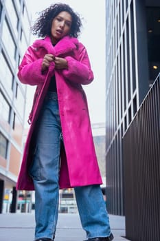 Vertical portrait of a fashionable young woman in a bold pink coat and jeans, walking confidently through a modern urban area. Her serious expression adds intensity to the stylish scene.