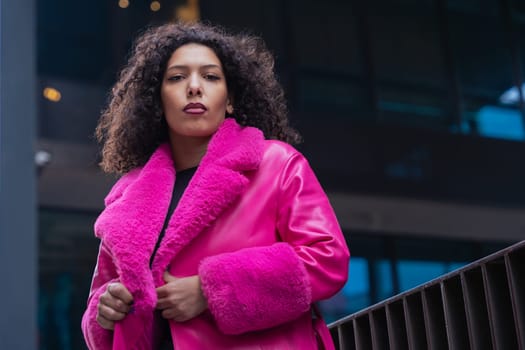 Horizontal portrait of a fashionable young woman in a bold pink coat and jeans, walking confidently through a modern urban area. Her serious expression adds looking at camera