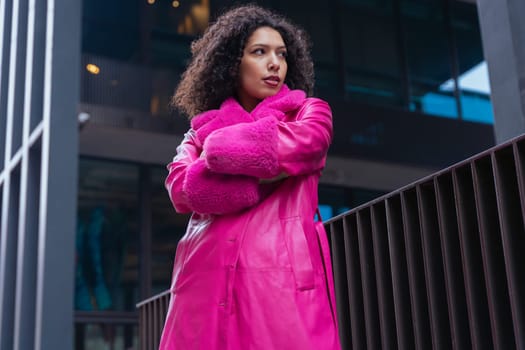 African american woman wearing a vibrant pink coat stands with arms crossed in a modern city setting. Her confident expression and bold fashion create a striking look against urban architecture.