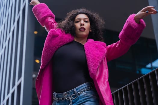 Graceful dancer with curly hair wearing a pink coat, expressing movement and energy in an urban environment. Modern fashion, contemporary dance, and artistic performance against a city backdrop.