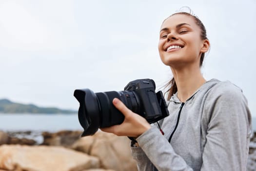 A joyful young female photographer in a gray hoodie, standing by the sea, capturing moments with her camera The image showcases happiness, creativity, and outdoor photography