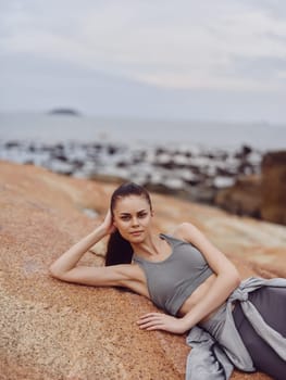 A young woman dressed in sporty activewear relaxes on a rock at the beach, embodying a healthy lifestyle and tranquility with a coastal backdrop