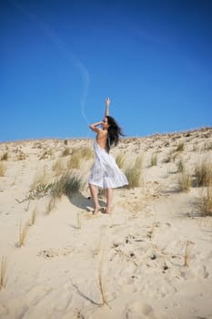 A stylish woman poses on a golden dune in a breezy summer dress. Her carefree posture and windblown hair highlight effortless beach fashion.
