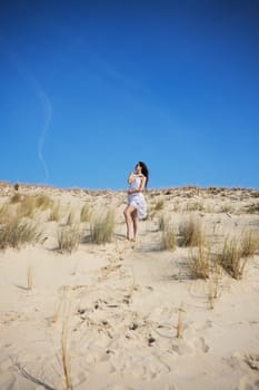 A lady in a flowy white dress explores a dry sand dune with wild grass, framed by a vivid blue sky, symbolizing adventure and harmony with nature