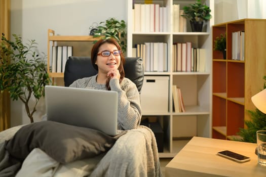 Cheerful woman wearing glasses sitting in an armchair with a laptop.