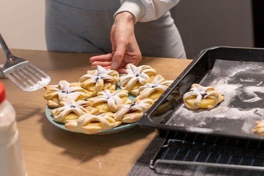 A woman pick the fruit puff pastries with spatula from baking tray and places them on ceramic plate.